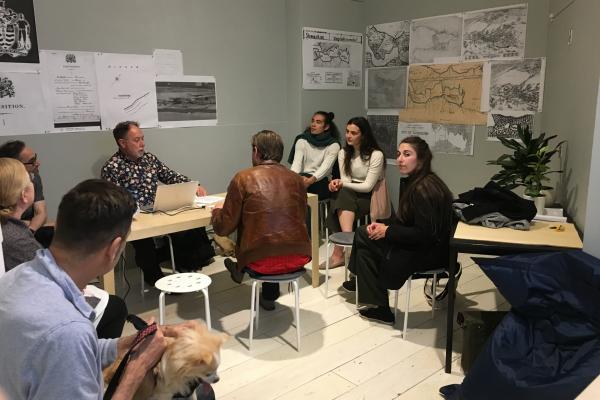 Gordon Brent Brochu Ingram sitting at a table with his computer talking to a room of about 8 people and a small dog during the 'ecological breakdown cabaret' performance event. They are sitting in the PLOT space at Access, with grey walls covered by historical maps printed out black and white.