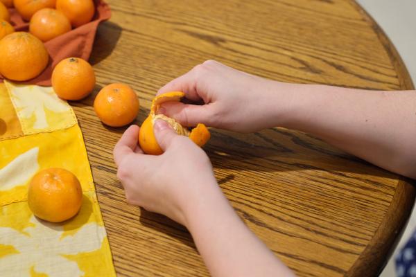 Two hands peeling a tangerine at a wooden table. On the left we see a corner of a bright yellow Bojagi (traditional Korean wrapping cloth), and a number of bright orange tangerines.