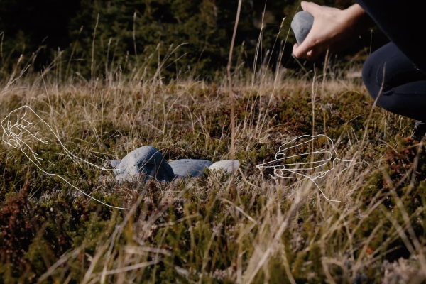 worm's eye view of a person's hands holding a large stone, crouched above a few stones in the grass. The image is overlaid with a white line drawing of hands beading.