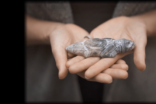 A still from Spirit Emulsion, showing artist Siku Allooloo's hands cradling a small reptile figurine carved from rock. The details are finely etched, with a small divot in the creature's back