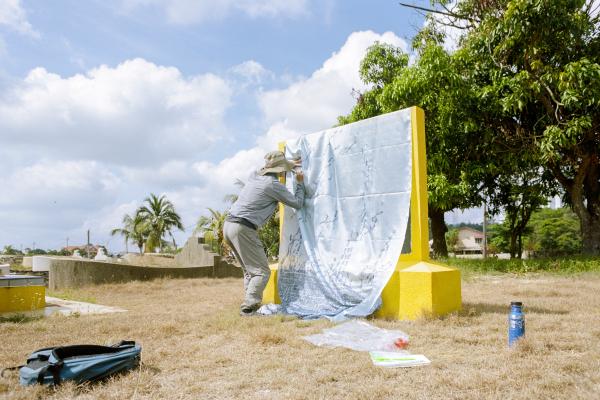 A photo of Alvin Luong working on a frottage. Fabric is draped over a bright yellow monument in front of Alvin, and he is wearing a sun hat and long sleeves. The grass beneath the monument is dry. 