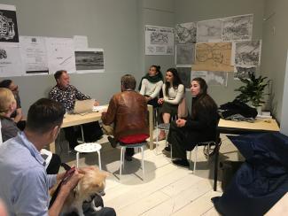 Gordon Brent Brochu Ingram sitting at a table with his computer talking to a room of about 8 people and a small dog during the 'ecological breakdown cabaret' performance event. They are sitting in the PLOT space at Access, with grey walls covered by historical maps printed out black and white.