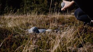 worm's eye view of a person's hands holding a large stone, crouched above a few stones in the grass. The image is overlaid with a white line drawing of hands beading.