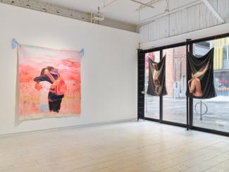 A corner of a gallery where the wall meets the window. On the left there is a painting of two figures embracing, to the right are two hanging pieces of photography on printed fabric. The first is a hand holding a crown, the other is a figure turned away holding a fan. 