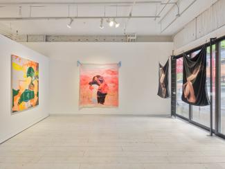 A view of a section of the gallery. On one wall there is a painting of a figure with overlaying elements of hopscotch. The wall across from the camera has a painting of two figures embracing. The wall to the right is mostly windows, with two hanging pieces. These are photographs printed on fabric, one is of a hand holding a microphone and the other is a figure turned away holding a fan. 