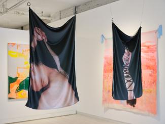 Two hanging photographs printed on fabric. The one closer is of a figure scratching themselves with a stick, the other is of a hand holding a microphone. 