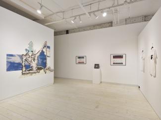 A corner of a gallery. On the right is a drawing of a figure emerging from a hole, there are elements of a cyanotype photograph surrounding it. The other wall are maps with printed elements overtop of them, and a beaded medalion in a shadowbox on a plinth. 