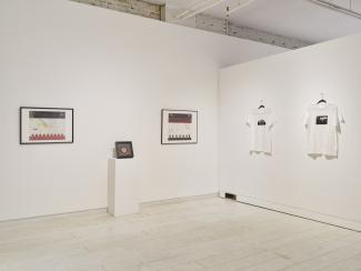 A corner of the gallery, on one wall there is maps with printed elements overtop of them, in the middle of these maps is a beaded medallion in a shadowbox on a plinth, the other wall has two hanging tshirts. 