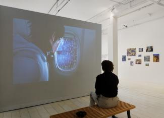 A person sits on a wooden bench, viewing a large video projection of a character looking out an airplane window. Paintings hang on the white gallery walls.