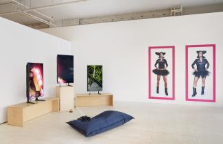 Art gallery with three vertical screens showing vibrant images, two large photos of two figures in a tutu on the wall, and a cushion with headphones in the centre. 
