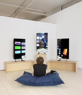 A person sits on a blue cushion wearing headphones, facing three digital screens displaying vibrant visuals from Sondi’s art installation piece on the display monitors.