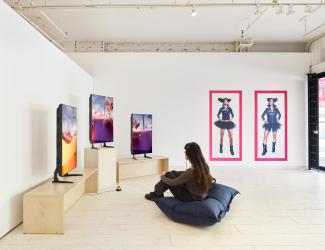 A person with long dark hair sits on a cushion wearing headphones, viewing three screens displaying ‘Home404’ by artist Sondi on three monitors. Two bold, colourful portraits hang on the wall to the left.