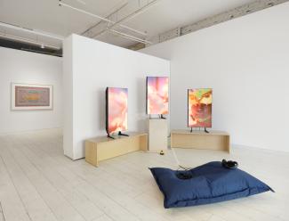​​Art installation with three vibrant digital screens displaying ‘Home404’ by artist Sondi. Wooden benches surround the screens, with a blue cushion and headphones on the floor.