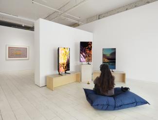 A person with long dark hair sits on a cushion wearing headphones, viewing three screens displaying ‘Home404’ by artist Sondi on three monitors.
