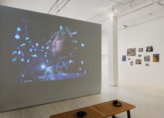  An immersive gallery setup featuring a large video projection of a person in a futuristic scene, surrounded by blue shards. A bench and headphones are in the foreground, with small colourful artworks displayed on a white wall to the right in the background.