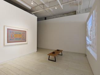 Gallery room featuring a colorful abstract painting on the left wall and a cityscape video projected on the right. A wooden gallery bench with headphones sits in the center.
