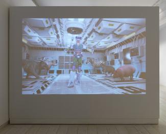 A futuristic scene is projected on a blank gallery wall, showing a person in colourful attire surrounded by a tiger, zebra, deer, and hippo inside a seemingly high-tech facility.