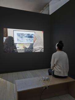 A projection plays a video work by Forensic Architecture. In the still image, an older man wears a keffiyeh, and points toward an aerial photograph. Captions read: "Some got killed (at the market), because they were there and did not go to the mosque."
