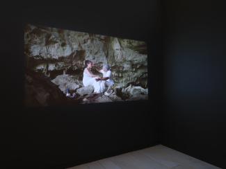 A projection still showing an older man, dressed in white, reaching towards the forehead of a dark-haired woman. They are sitting in a large rock cavern. The woman has red paint marking her. 