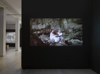 A projection still showing an older man, dressed in white, reaching towards the forehead of a dark-haired woman. They are sitting in a large rock cavern. There is a second video projection showing a flower visible in the background of the gallery. 