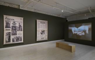 Two large newspaper enlargements stretch from floor to ceiling, each with spreads from "Indígena: News From Indian America." A projection on another wall shows two hands cradling a carved stone figure.